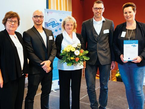 Links steht die Ministerin für Arbeit, Soziales und Integration Sachsen-Anhalt. Rechts neben ihr stehen 4 Mitarbeiter und Mitarbeiterinnen von RM Balance. Sie freuen sich und haben Blumen und eine Urkunde in der Hand. Alle schauen in die Kamera.