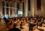 Viele Menschen sitzen bei einer Festveranstaltung in einer Kirche. Auf der Bühne stehen Redner:innen.