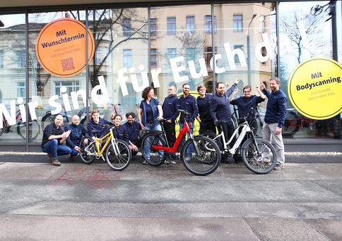 12 Menschen stehen mit 3 Fahrrädern vor einem großen Schaufenster.