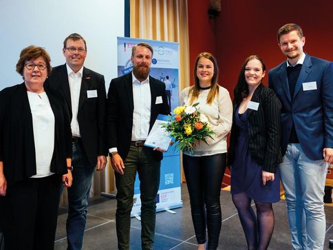 Die Minsiterin für Arbeit, Soziales und Integration steht links. Rechts neben ihr stehen 5 Mitarbeiter und Mitarbeiterinnen von Umland mit Blumen und einer Urkunde. Alle freuen sich und schauen in die Kamera.