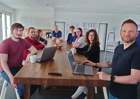 Mehrere Personen stehen an einem großen Tisch. Auf dem Tisch sind zwei Laptops und einige Tassen