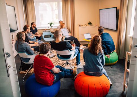 Mehrere Personen sitzen auf Stühlen und Sitzsäcken in einem Raum und schauen auf einen großen Bildschirm an der Wand